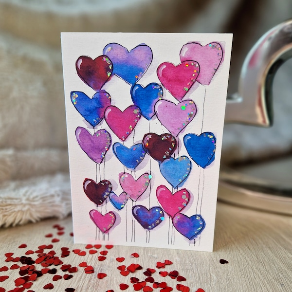 Puede incluir: Una pintura de acuarela de una tarjeta con un grupo de globos en forma de corazón rosa, morado y azul. Los corazones están pintados con purpurina y tienen cuerdas atadas a ellos.