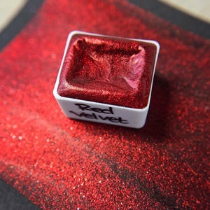 Könnte beinhalten: Ein quadratischer Aquarellfarbkasten gefüllt mit schimmernder roter Farbe, beschriftet mit "Red Velvet" in einem weißen Behälter. Die Farbe liegt auf einer glitzernden roten Oberfläche mit schwarzem Rand.