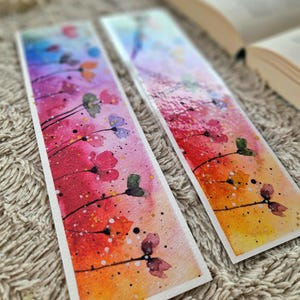 May include: Two watercolor bookmarks with a colorful floral design. The bookmarks feature a gradient background with red, orange, yellow, and blue hues. The flowers are painted in shades of pink, purple, and green. The bookmarks are perfect for adding a touch of color and whimsy to any book.