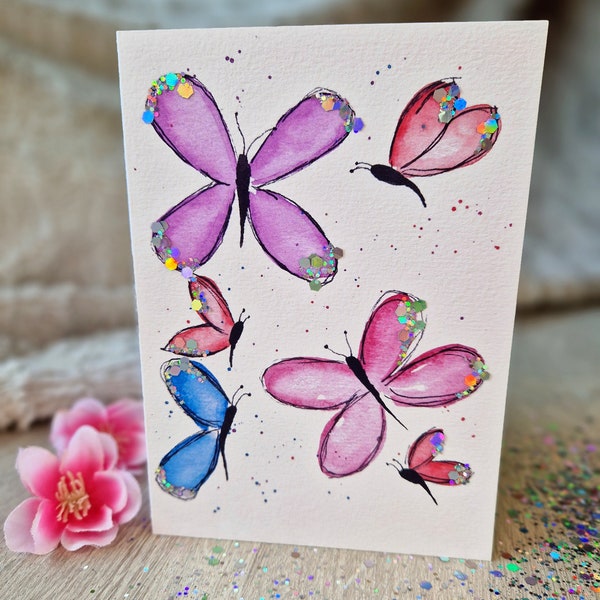 Puede incluir: Una tarjeta blanca con una pintura de acuarela de cinco mariposas. Las mariposas son rosas, azules y moradas con detalles de purpurina.