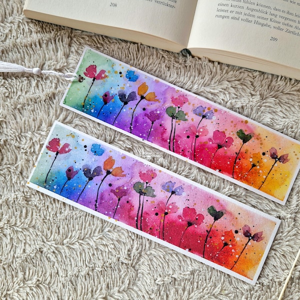 Puede incluir: Dos marcadores de acuarela con un fondo degradado de arcoíris y flores de colores. Los marcadores son blancos con un borla.