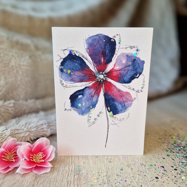 Puede incluir: Una pintura de acuarela de una flor azul y roja con detalles de purpurina en una tarjeta blanca.