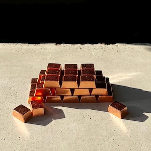 Puede incluir: Una pila de pequeñas barras de color cobre dispuestas en forma de pirámide. Las barras están esparcidas sobre una superficie gris claro.