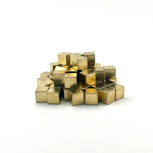 May include: A collection of small, gold-coloured metal cubes. The cubes are arranged in a pile, creating a textured surface. The lighting reflects off the edges and surfaces, giving them a shiny appearance.