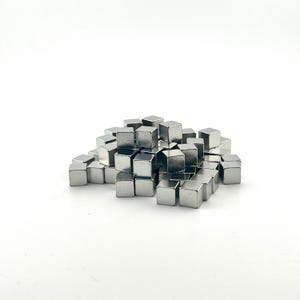 Könnte beinhalten: Ein Haufen kleiner, glänzender, silberfarbener Metallwürfel. Die Würfel sind gleichmäßig geformt und scheinen aus einem reflektierenden Material zu bestehen. Sie sind auf einer weißen Oberfläche verstreut und bilden ein geometrisches Muster.