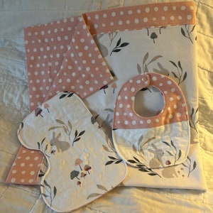 May include: A set of three baby items in a woodland theme. A pink and white bib with white polka dots, a pink and white burp cloth with white polka dots, and a white burp cloth with a woodland print of mushrooms, leaves, and bunnies.