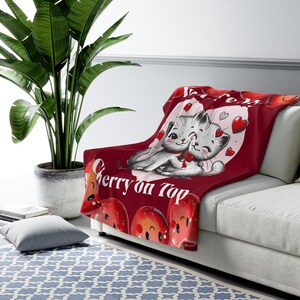Puede incluir: Manta de forro polar roja con un corazón blanco y dos gatos de dibujos animados en su interior. Los gatos se abrazan y llevan lazos rojos. El texto "We're a Cherry on Top" está impreso debajo del corazón.