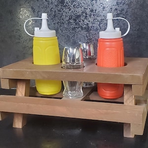 Pode incluir: Um suporte para condimentos de madeira em forma de mesa de piquenique com duas garrafas de pressão, uma amarela e uma laranja, e dois agitadores de vidro transparentes.