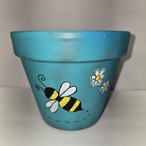 May include: A turquoise ceramic flower pot with a hand-painted design. The pot features a black and yellow bee with white wings and two small white daisies. The pot is designed for planting flowers and other plants.