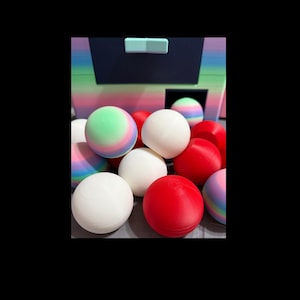 May include: A collection of colorful balls, including white, red, and pastel rainbow-colored spheres. The balls are in front of a dark blue structure with a light blue handle. The balls appear to be 2.5 inches in diameter.