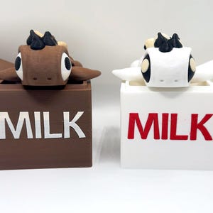 May include: Two milk cartons with cow figurines on top. The carton on the left is brown with white text that says "MILK". The carton on the right is white with red text that says "MILK".