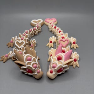 May include: Two small, hand-painted ceramic dragons facing each other. The dragons are decorated with white and pink icing, and have red eyes. The dragons are in a heart shape.