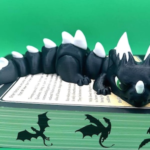 May include: A black 3D printed dragon bookmark with white spines and green eyes. The dragon is curled up on a book with other dragon silhouettes printed on the cover.