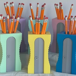 May include: Five colourful pencil sharpeners with different coloured pencils in each. The sharpeners are made of plastic and have a ribbed texture. Each sharpener has a grey metal blade and a small hole for sharpening pencils. The pencils are all different colours, including red, blue, green, yellow, and orange.