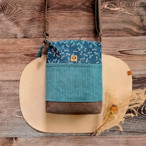 Könnte beinhalten: Eine Umhängetasche aus türkisfarbenem Cord mit einem blauen Libellenmuster oben und einem braunen Boden. Die Tasche hat einen grauen Riemen und eine kleine Quaste. Ein kleines Holzschild befindet sich auf der Vorderseite.