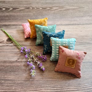 Könnte beinhalten: Eine Sammlung kleiner, quadratischer Zierkissen in verschiedenen Farben, darunter Rosa, Gelb und Türkis, mit Libellen- und Blumenmustern. Ein Zweig Lavendel liegt vor den Kissen. Die Kissen haben ein aufgenähtes Lederetikett.