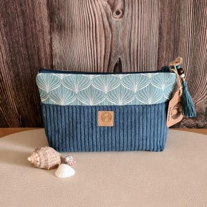 Könnte beinhalten: Eine blaue Kosmetiktasche aus Cord mit einem türkis-weiß gemusterten Oberteil. Die Tasche hat einen Reißverschluss, eine Quaste und einen kleinen Lederaufnäher. Muscheln sind vor der Tasche angeordnet.