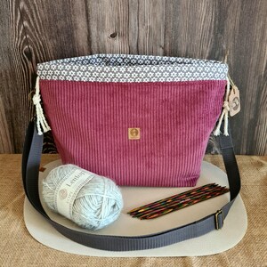 Könnte beinhalten: Burgunderfarbene Cord-Tragetasche mit grauem, verstellbarem Riemen. Die Tasche hat ein grau-weißes Blumenmusterfutter. Ein Knäuel hellblauer Wolle und ein Satz Stricknadeln aus Holz liegen neben der Tasche.