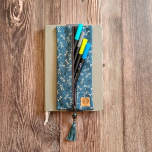 Könnte beinhalten: Ein tealfarbenes Federmäppchen mit Libellenmuster, geöffnet, um farbige Marker zu zeigen. Das Etui hat einen Reißverschluss und eine Quaste. Es liegt auf einem grauen Notizbuch, alles auf einer Holzoberfläche.