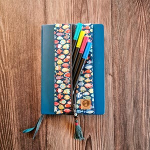 Könnte beinhalten: Ein blaues Notizbuch mit einem Federmäppchen mit bunten Fischen. Das Federmäppchen ist geöffnet und zeigt mehrere farbige Marker. Ein Quaste hängt am Reißverschluss. Auf einer Holzoberfläche.