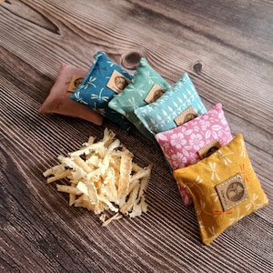 Könnte beinhalten: Eine Auswahl an kleinen, quadratischen Stoffsäckchen mit verschiedenen Mustern, darunter Libellen und Blumen. Jedes Säckchen hat ein Lederetikett mit einem Logo. Daneben liegt ein Haufen hellfarbiger Holzspäne.