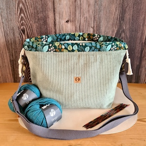 Könnte beinhalten: Eine hell salbeigrüne Cord-Projekttasche mit Kordelzug und einem Innenfutter mit dunkelgrünem Blumenmuster. Die Tasche verfügt über einen grauen, verstellbaren Schultergurt, ein kleines quadratisches Lederlogo, zwei Knäuel blaugrünes Garn und Stricknadeln.