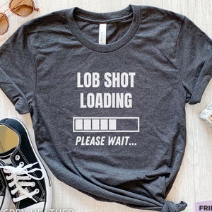 May include: A dark grey heather short-sleeve t-shirt with white text that reads LOB SHOT LOADING and PLEASE WAIT, featuring a white loading bar graphic. The shirt is displayed with black and white trainers and brown-tinted sunglasses.