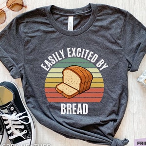 Puede incluir: Una camiseta gris jaspeado oscuro con un gráfico de una hogaza de pan con una rebanada delante, sobre un fondo circular de estilo retro de puesta de sol con rayas horizontales en verde, amarillo y naranja, y el texto EASILY EXCITED BY BREAD.