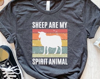 Puede incluir: Una camiseta gris oscuro jaspeado con la silueta blanca de una oveja centrada sobre un gráfico de rayas horizontales desgastadas en rojo, naranja, amarillo y turquesa. El texto "SHEEP ARE MY SPIRIT ANIMAL" está impreso en blanco sobre el gráfico.