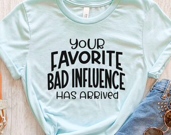 Puede incluir: Una camiseta de manga corta de color azul claro con texto negro impreso en la parte delantera que dice: YOUR FAVORITE BAD INFLUENCE HAS ARRIVED. La camiseta cuenta con un cuello redondo y está hecha de una tela suave, en color azul hielo.