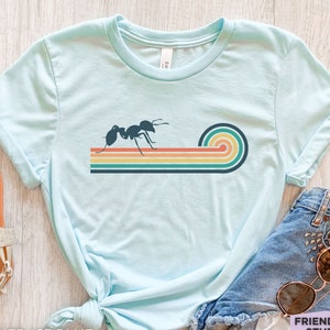 May include: A light blue t-shirt featuring a dark blue ant graphic walking along a series of horizontal, multi-coloured stripes that curve upwards at the end. The stripes are coloured in shades of orange, yellow, teal, and dark blue.