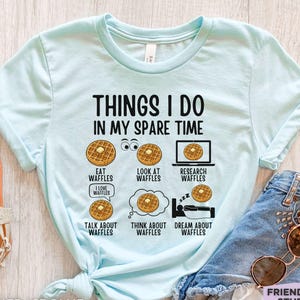 May include: A light blue t-shirt featuring the black text "THINGS I DO IN MY SPARE TIME" above six illustrations of waffles with butter. The graphics include labels for eating, looking at, researching, talking about, thinking about, and dreaming about waffles.