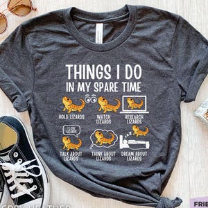 Può includere: Una t-shirt grigio scuro mélange con il testo bianco THINGS I DO IN MY SPARE TIME e sei illustrazioni di draghi barbuti con le etichette: HOLD LIZARDS, WATCH LIZARDS, RESEARCH LIZARDS, TALK ABOUT LIZARDS, THINK ABOUT LIZARDS e DREAM ABOUT LIZARDS.