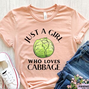 Könnte beinhalten: Ein pfirsichfarbenes Kurzarm-T-Shirt mit einer Grafik eines grünen Kohls in der Mitte. Der Text "JUST A GIRL WHO LOVES CABBAGE" ist in schwarzer Schrift über und unter der Kohl-Illustration auf der Vorderseite des T-Shirts aufgedruckt.