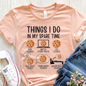 以下が含まれることがあります： 「THINGS I DO IN MY SPARE TIME」の文字とクッキーのイラストが描かれた、ピーチ色のTシャツ。Tシャツには「BAKE COOKIES」、「WATCH COOKIE VIDEOS」、「EAT COOKIES」、「TALK ABOUT COOKIES」、「THINK ABOUT COOKIES」、そして「DREAM ABOUT COOKIES」のフレーズも含まれています。