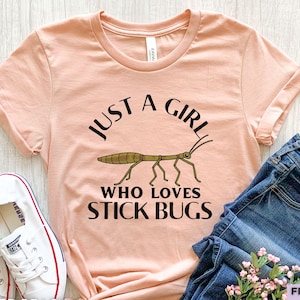May include: A peach-coloured short-sleeve t-shirt featuring a graphic of a brown stick bug in the centre. The text printed in black above and below the insect reads, JUST A GIRL WHO LOVES STICK BUGS. The shirt is paired with blue denim jeans.