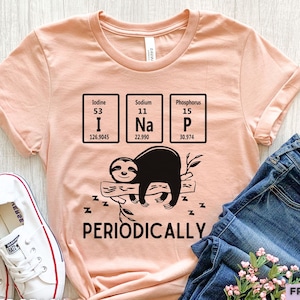 Puede incluir: Una camiseta de manga corta color melocotón con un gráfico negro de un perezoso durmiendo en una rama. Sobre el perezoso hay tres cuadros de la tabla periódica para yodo, sodio y fósforo, que deletrean I Na P. Debajo del perezoso está el texto PERIODICALLY.