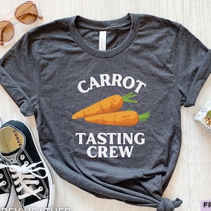 May include: A dark grey heather t-shirt featuring a graphic of two orange carrots with green tops. The text above the carrots reads CARROT and the text below reads TASTING CREW in white capital letters. The shirt has short sleeves and a crew neckline.