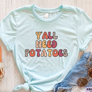 May include: A light blue short-sleeve t-shirt featuring the text Y'ALL NEED POTATOES in a multi-colored, rounded, retro-style font. The shirt is displayed alongside a pair of light-colored sandals, denim shorts, and sunglasses on a light surface.