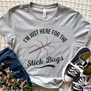 May include: A light grey short-sleeve t-shirt featuring a brown stick bug illustration centred between the black text that reads I'M JUST HERE FOR THE Stick Bugs. The shirt is paired with dark blue denim jeans and black and white trainers.