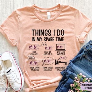Könnte beinhalten: Ein pfirsichfarbenes T-Shirt mit dem Text "Things I do in my spare time" und Illustrationen von Axolotln. Der Text unter den Illustrationen umfasst "Feed axolotls", "Look at axolotls", "Research axolotls", "Talk about axolotls", "Think about axolotls" und "Dream about axolotls".