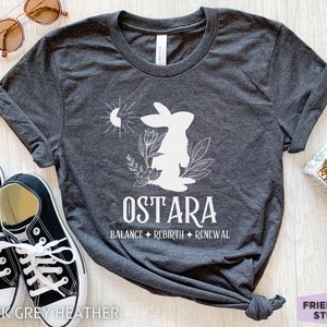 Puede incluir: Una camiseta gris jaspeado oscuro con un gr&aacute;fico blanco de un conejo, sol, luna y follaje. El texto dice OSTARA, BALANCE, REBIRTH y RENEWAL. La prenda tiene mangas cortas, cuello redondo y una apariencia de tejido suave y texturizado.