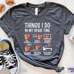 May include: Dark grey heather t-shirt with a white graphic that says "Things I do in my spare time" and features a series of illustrations of squirrels. The illustrations depict squirrels feeding, looking at a computer, researching, talking, thinking, and sleeping.