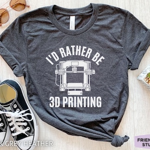 以下が含まれることがあります： 濃いグレーのヘザーTシャツに、白い文字で「3Dプリントする方が好き」と書かれています。テキストの上に、白い輪郭の3Dプリンターがあります。