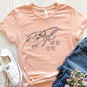 May include: A peach-coloured t-shirt featuring a black lobster illustration and the text "SAY NO TO POT." The shirt is styled with blue jeans and white trainers. The image is from Friend Tee Studio.