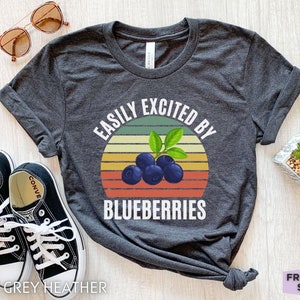 Op de afbeelding: Donkergrijs gemêleerd T-shirt met de witte tekst "EASILY EXCITED BY BLUEBERRIES", met een afbeelding van bosbessen en een retro zonsondergangontwerp. Het shirt wordt gecombineerd met zwarte sneakers en een zonnebril.