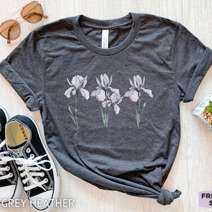 Peut inclure: Un t-shirt gris foncé chiné avec un motif de trois fleurs d'iris blanches avec des accents violets. Le t-shirt est posé à plat sur une surface en bois clair avec une paire de baskets noires et une petite plante en pot.