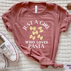 Könnte beinhalten: Ein heidefarbenes T-Shirt mit dem Text "Just a girl who loves pasta" und einer Grafik von Pasta. Das Shirt liegt flach auf einer weißen Oberfläche.