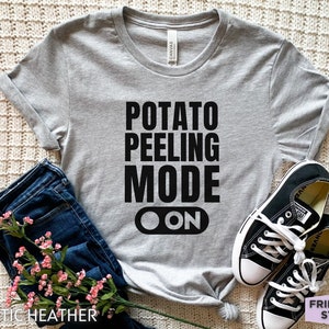 May include: A gray t-shirt with the text "POTATO PEELING MODE ON" in black. The shirt is paired with blue jeans and black sneakers. The words "ATHLETIC HEATHER" and "FRIEND TEE STUDIO" are also visible.