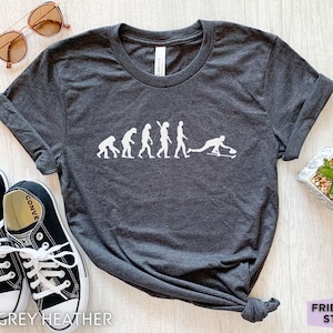 May include: A dark grey heather t-shirt with a white graphic of the evolution of man ending with a person playing curling. The text "FRIEND TEE STUDIO" is visible in the bottom right corner of the image.